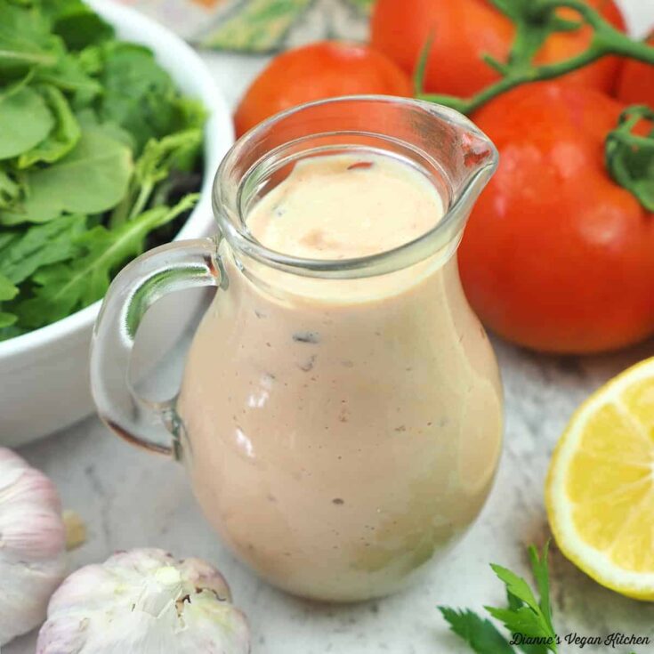Vegan salad dressing in a glass pitcher with fresh vegetables and lemon.