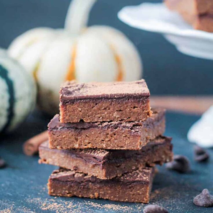 Vegan chocolate fudge squares stacked on a dark surface with pumpkins in the background.