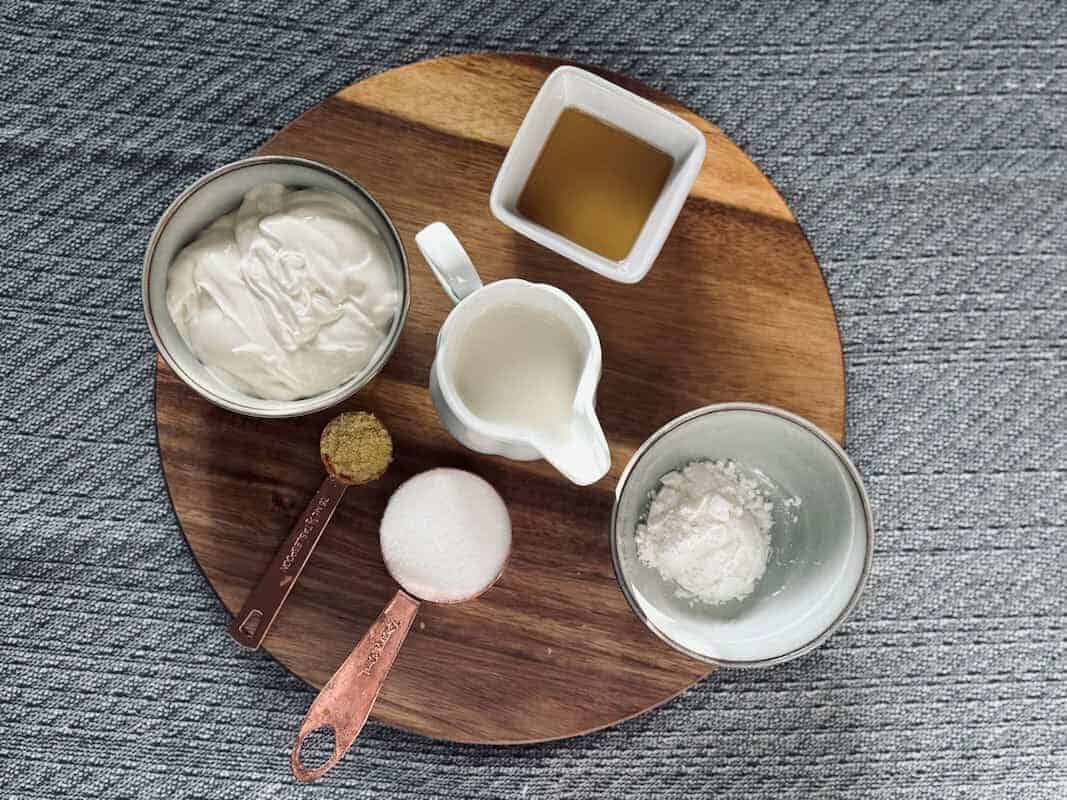 Vegan baking ingredients including flour, sugar, oil, and plant-based yogurt on a round wooden board.